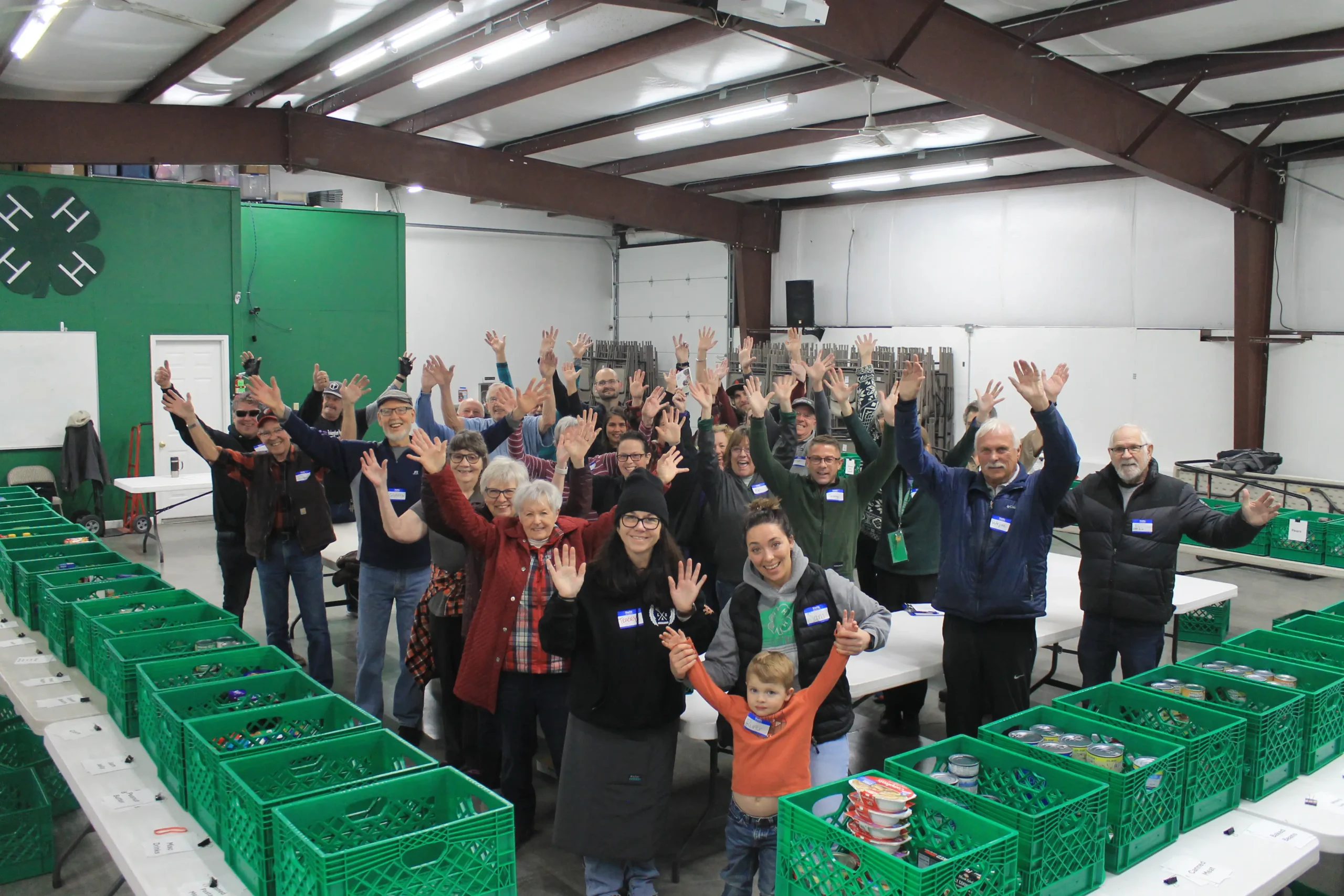 Volunteers smile and wave while standing behind rows of green donation bags and collection bins at a Crook County Green Bag collection day.