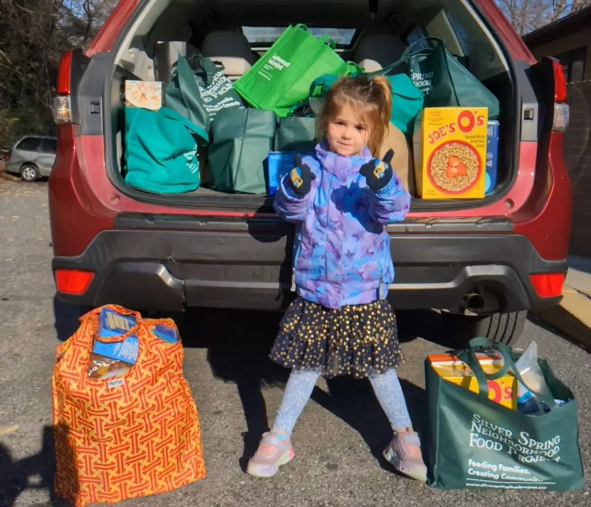Young girl gives two thumbs up beside an open SUV trunk packed with green Silver Spring Neighborhood Food Project donation bags